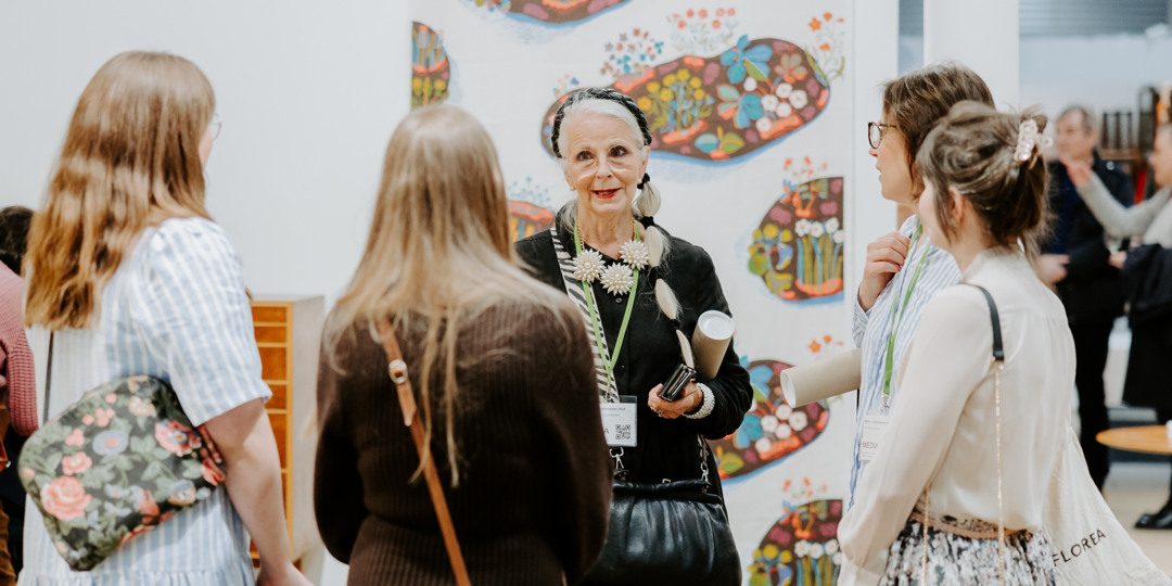 Mörkgrön sammetssoffa mot olivgrön vägg, med träbord bredvid som håller en glasvas med grenar och två koppar; rund jutematta i förgrunden.